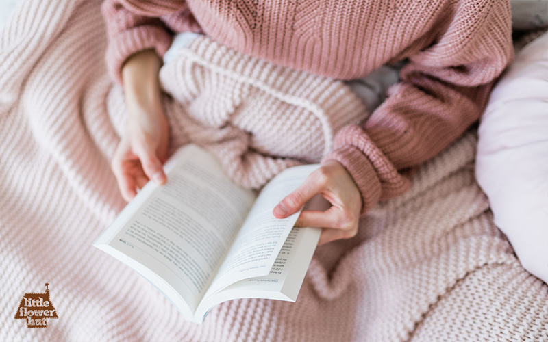 A lady wearing a pink shirt covering a pink blanket reading a book